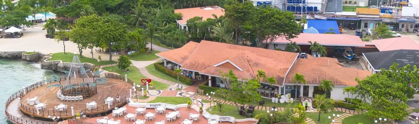 Aerial drone shot of Jpark Island Resort and Waterpark in Mactan, Cebu, showing the resort's coastline, buildings, and swimming pool complex under a blue sky.